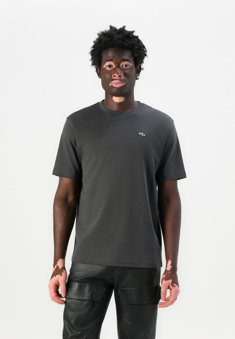 T-shirt gris foncé à manches courtes avec un col rond et un logo sur la poitrine, associé à un pantalon en cuir noir avec des poches cargo.