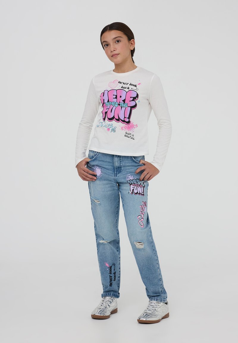 Camicia bianca a maniche lunghe con testo e grafiche colorate; jeans azzurri chiari con toppe e dettagli strappati sulla parte anteriore. Scarpe argentate.