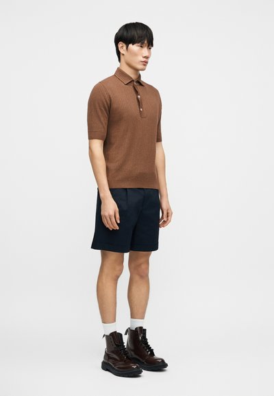 Polo marron côtelé à manches courtes avec patte de boutonnage à trois boutons, associé à un short bleu marine foncé. Le mannequin porte des bottes marron et des chaussettes blanches.