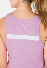 Donna con i capelli castano chiaro raccolti in una coda di cavallo, che indossa un top senza maniche rosa con una striscia orizzontale bianca sulla parte superiore della schiena.