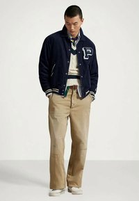 Νεαρός άνδρας που φορά ναυτικό varsity μπουφάν, κρεμ πουλόβερ με σταυρωτό πλέξιμο, ρίγες πουκάμισο, μπεζ παντελόνι και λευκά αθλητικά παπούτσια, στέκεται με τα χέρια στις τσέπες.