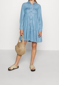 Denimkjoja me një përparëse me dük, dy xhepa në gjoks, fund të mbledhur, mëngë të gjata dhe ngjyrë të kaltër të lehtë. Çanta të endura dhe sandale bezhë.