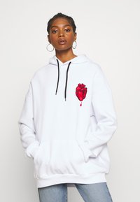 White oversized hoodie with black drawstrings and front pocket, featuring a red embroidered heart graphic on the chest. Fleece material.