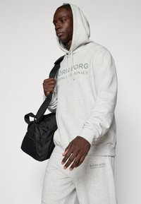 Γκρι φούτερ με κουκούλα και πράσινο κείμενο, ταιριαστά κολάν και μαύρη τσάντα. Διαθέτει μεγάλο κεντρικό λογότυπο και κουκούλα με κορδόνι.