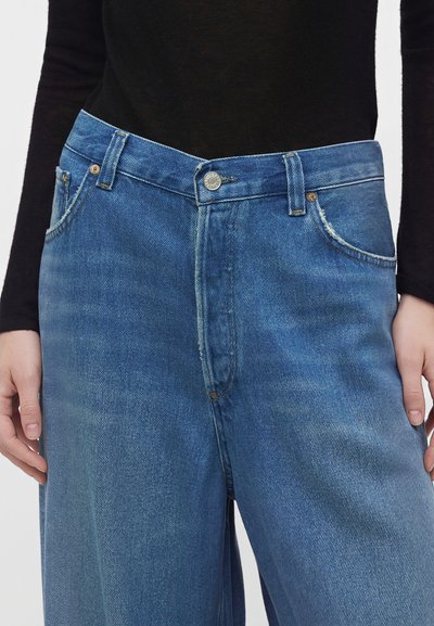 Personne portant un jean en denim bleu taille haute et ample, ainsi qu'un haut noir à manches longues ajusté avec les mains reposant le long du corps.