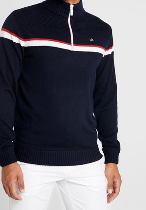 Maglione blu navy con mezza zip, con strisce rosse e bianche sul petto e logo "ck", indossato con pantaloni bianchi.