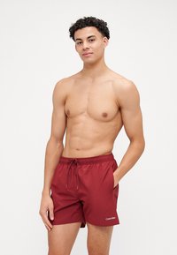 Shorts de bain bordeaux avec une taille élastique et un cordon de serrage, disposant de poches latérales et d'un logo Calvin Klein blanc. Texture lisse.