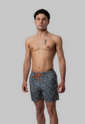 Giovane uomo in piedi senza maglietta, indossa pantaloncini da bagno fantasia con coulisse arancione e un tatuaggio verticale sul petto sinistro.