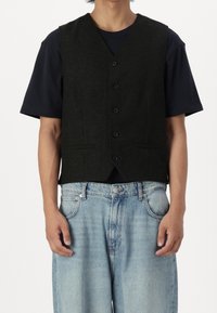 Gilet nero in lana con scollo a V, chiusura con cinque bottoni e tasche laterali, indossato sopra una t-shirt blu navy, abbinato a jeans larghi azzurri.