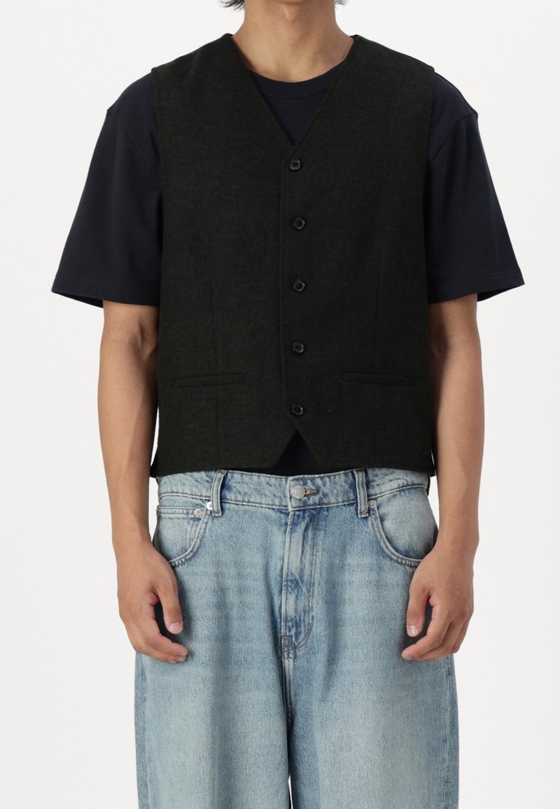 Gilet nero in lana con scollo a V, chiusura con cinque bottoni e tasche laterali, indossato sopra una t-shirt blu navy, abbinato a jeans larghi azzurri.