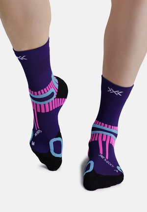 Voeten met paarse, roze, blauwe en zwarte sportsokken met geometrische patronen, gezien van achteren en iets van boven tegen een witte achtergrond.