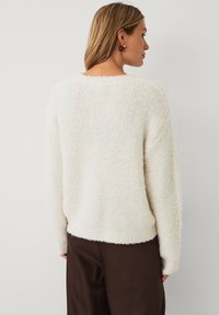 Kvinde med skulderlangt blont hår iført en hvid, fuzzy sweater og mørkebrune bukser, som står vendt væk mod en ensfarvet baggrund.