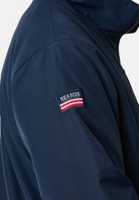 Mørkeblå jakke med en glat tekstur. Har en sidelomme med lynlås og et patch med "SEASIDE" samt røde og hvide striber på ærmet.