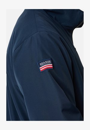 Granatowa kurtka o gładkiej teksturze. Posiada boczną kieszeń na zamek oraz naszywkę z napisem "SEASIDE" oraz czerwono-białymi paskami na rękawie.