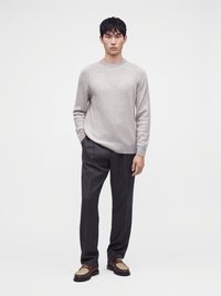 Homme portant un pull gris clair, un pantalon plissé noir et des mocassins marron bicolores, debout devant un fond blanc uni.