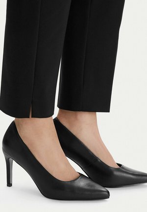 Mujer con zapatos de tacón negros de punta afilada combinados con pantalones negros a medida de longitud hasta el tobillo sobre un fondo blanco.