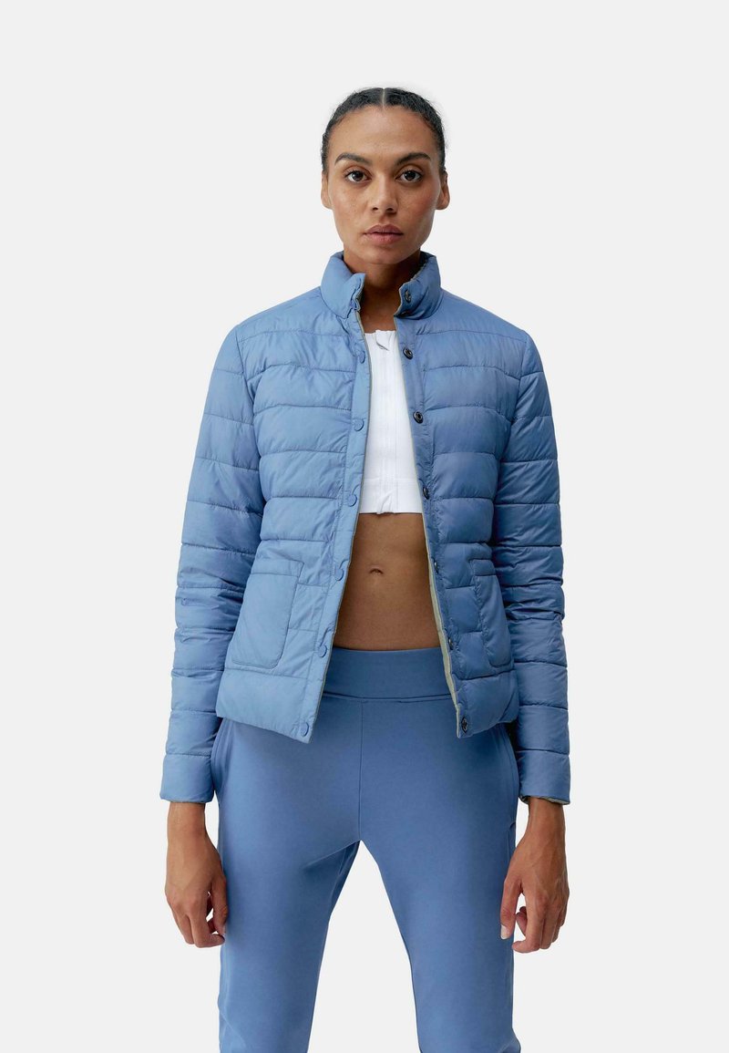 Lichtblauwe geïsoleerde jas met een staande kraag, gekruiste textuur, enkele knoopsluiting en zijzakken. Draagt over een witte top.