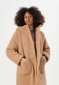 Übergroßer beigefarbener Fleecepullover mit Kapuze, strukturiertem Oberflächenmaterial, Seitentaschen und gerippten Strickmanschetten. Glattes Innenfutter, das von innen sichtbar ist.