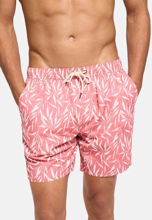 Hombre con pantalones cortos de baño rosa con patrón de hoja tropical blanca y cordón, con las manos en los bolsillos, sobre un fondo liso.
