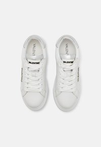 White leather sneakers with a smooth texture, round toe, and silver accents. Features contrasting black logo and fabric laces.