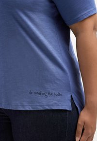 Blauwe T-shirt met korte mouwen, gemaakt van een zachte stof en met zijsplitsen. Heeft zwarte tekst aan de onderkant: "doe vandaag iets blauws..."