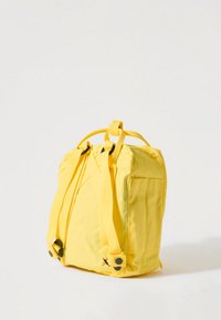 Dzeltena auduma mugursoma ar regulējamām siksnām, taisnstūrveida forma, priekšējā kabata, gluda tekstūra un pastiprinātas šūšanas detaļas.