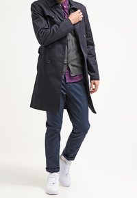 Dunkelblauer, doppelt geknöpfter Mantel über einer grauen Weste, getragen mit einem karierten Hemd und marineblauen Hosen. Ergänzt durch weiße Turnschuhe.