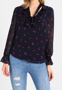 Donna che indossa una blusa trasparente blu navy con stampa di ciliegie rosse, colletto a fiocco, maniche con volant, abbinata a jeans strappati blu chiaro.