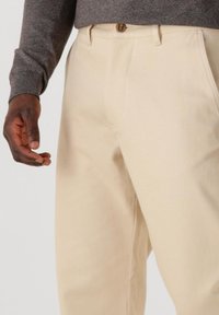 Bež pamuk hlače s kopčanjem na dugme, vidljivim bočnim džepovima i opuštenim krojem, uparen s tamno sivim puloverom.