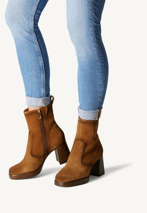 Bruna mocka ankboots med klack och dragkedja på sidan, i kombination med ljusblå uppvikta jeans, som visar en texturerad yta och strömlinjeformat design.