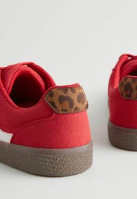 Czerwone zamszowe sneakersy z teksturowanym brązowym akcentem w zwierzęcy wzór na pięcie oraz formowaną brązową gumową podeszwą, z białym pasem po boku.