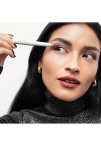 Crayon à yeux blanc avec un embout violet, tenu près de l'œil. Le sujet porte un col roulé noir pailleté et des boucles d'oreilles dorées. Peau lisse et maquillage neutre.