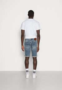 Man som vänder bort ansiktet, iklädd en vit t-shirt, uppvikta denimshorts, vita strumpor med svarta ränder och vita sneakers, mot enfärgad bakgrund.