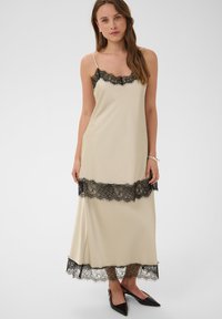 Femme portant une robe longue beige sans manches avec un bord en dentelle noire au niveau de l'encolure, de la taille et de l'ourlet, associée à des ballerines noires pointues.