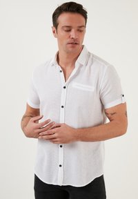 Camisa blanca de manga corta en tejido texturizado con botones negros y un bolsillo en el pecho. Presenta mangas enrolladas y un dobladillo curvado.