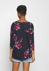 Maglietta floreale blu navy con maniche a tre quarti, caratterizzata da motivi di rose rosse e bianche. Tessuto morbido con taglio rilassato e scollatura rotonda.