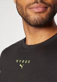 Černé sportovní tričko s kulatým výstřihem, vyrobené z hladké tkaniny, s nápisem "HYROX" a logem Puma ve výrazně žluté barvě na hrudi.