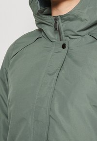 Green insulated jacket featuring a hood, zip closure, snap button, and contrasting stitching on smooth, matte fabric.