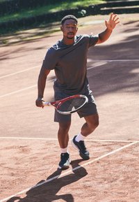 Mann i idrettsbekledning som spiller tennis på grusbane, holder racketen og forbereder seg på å treffe ballen med en backhand-sving.