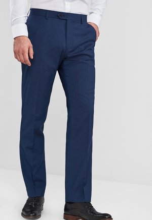 Mężczyzna ubrany w granatowe spodnie garniturowe, białą koszulę i czarne buty brogues stoi z jedną ręką w kieszeni na jednolitym tle.
