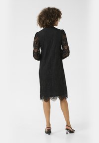 Black lace dress with long, puffed sleeves and scalloped hem. Intricate floral pattern throughout. Model wears black heeled sandals.