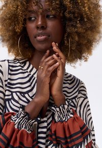 Koszula z kołnierzykiem w czarno-białym paskowanym wzorze, z obszerna czerwono-czarnymi rękawami i elastycznymi mankietami. Widoczne złote kolczyki w kształcie kół.