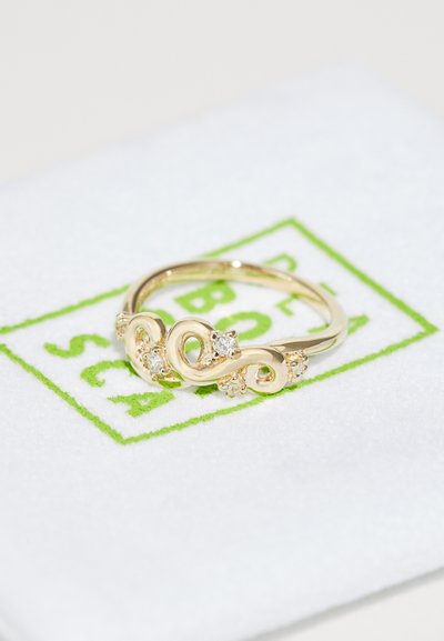 Anillo de oro con un diseño intrincado en espiral, que cuenta con pequeñas piedras preciosas transparentes engastadas en la parte frontal. Exhibido sobre un paño blanco con texto verde.