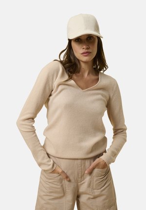 Maglione beige a maglia con scollo a V e maniche lunghe, abbinato a pantaloni cargo beige chiaro. Il modello indossa anche un cappello a coste color crema.