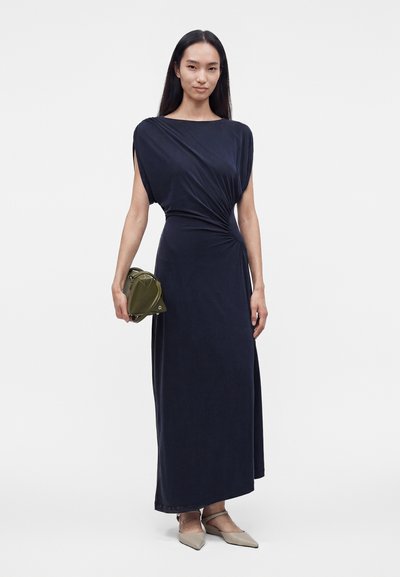 Femme debout, portant une robe bleu marine drapée, tenant un sac à main vert olive et des chaussures beiges pointues, sur fond blanc uni.