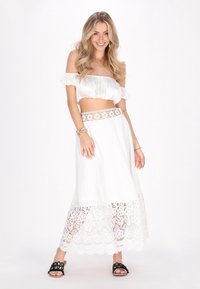 Weißes, schulterfreies Crop-Top mit puffärmel, kombiniert mit einem langen weißen Rock mit gewelltem Spitzenrand und floralem Akzent an der Taille.