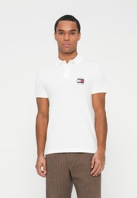 Mies, joka pukeutuu valkoiseen Tommy Hilfiger -pikeepaitaan, jossa on logo, ja ruskeisiin tekstiilipohjaisiin housuihin, seisoo yksinkertaista valkoista taustaa vasten.