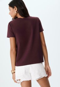 T-shirt corto color marrone bordeaux realizzato in tessuto morbido, con collo a giro e vestibilità comoda, abbinato a pantaloni corti in denim bianco sbiadito.