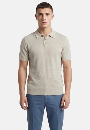 Junger Mann mit kurzem dunklem Haar, der ein beiges Strickhemd mit kurzen Ärmeln und Kragen sowie blaue Hosen trägt und vor einem schlichten weißen Hintergrund steht.