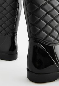 Bottes de pluie noires brillantes avec un motif matelassé sur la partie supérieure, texture lisse et bout arrondi. Semelle en caoutchouc basse avec légère traction.
